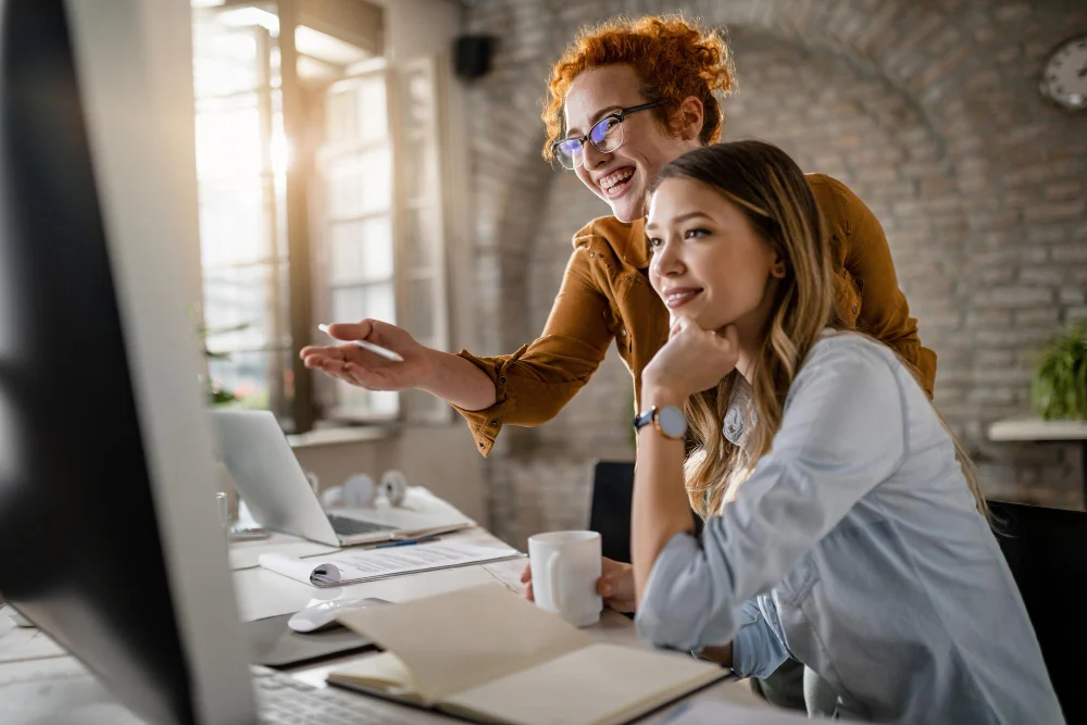 Dia do trabalhador: Empresárias felizes lendo um e-mail no computador enquanto trabalham juntas no escritório